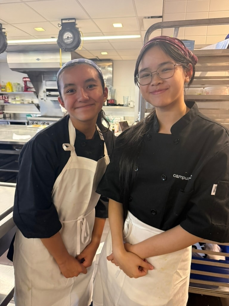 Two Summer Culinary students in chef jackets and aprons smiling in the kitchen